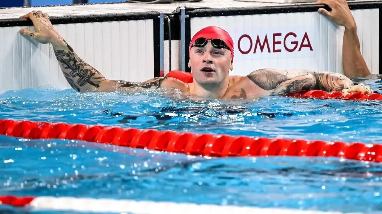 Adam Peaty, con cuffia rossa e occhialini, emerge dall'acqua della piscina, aggrappato al bordo.