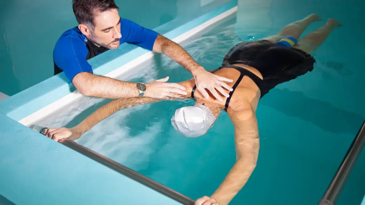 Fisioterapista assiste donna in piscina per ginnastica in acqua per mal di schiena. Lei galleggia con un supporto, lui la guida.