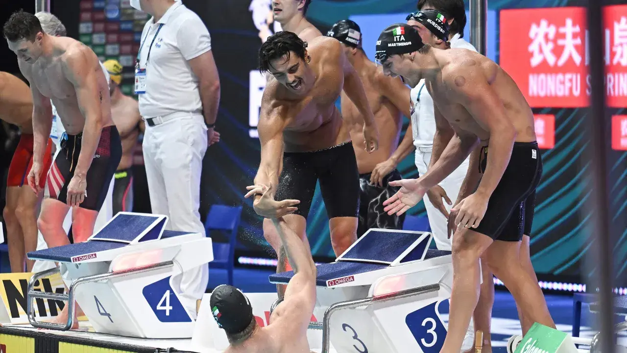 Atleti italiani festeggiano dopo la staffetta 4x100. Un nuotatore porge la mano a un compagno, gioia e sudore bagnano la corsia.