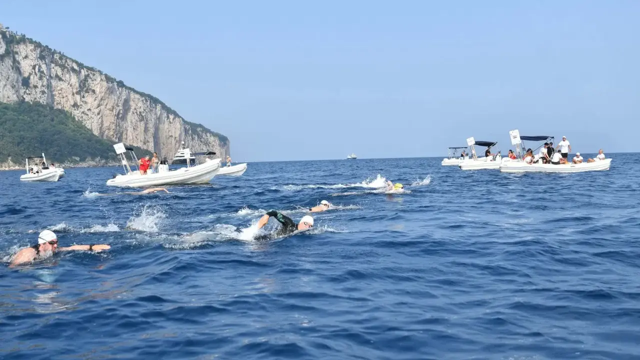 Nuotatori in gara tra Capri e Napoli, con barche di supporto e la costa rocciosa sullo sfondo.