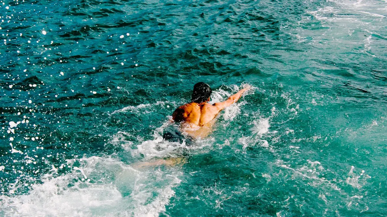 Uomo con schiena muscolosa nuotare in mare aperto, circondato da spruzzi d'acqua cristallina.
