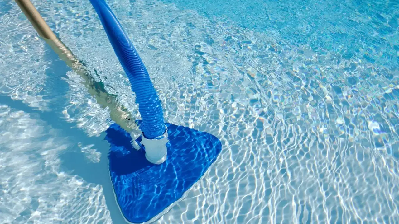 Pulizia di una piscina sporca con aspiratore blu e manico in legno. L'acqua limpida riflette la luce creando un effetto scintillante.