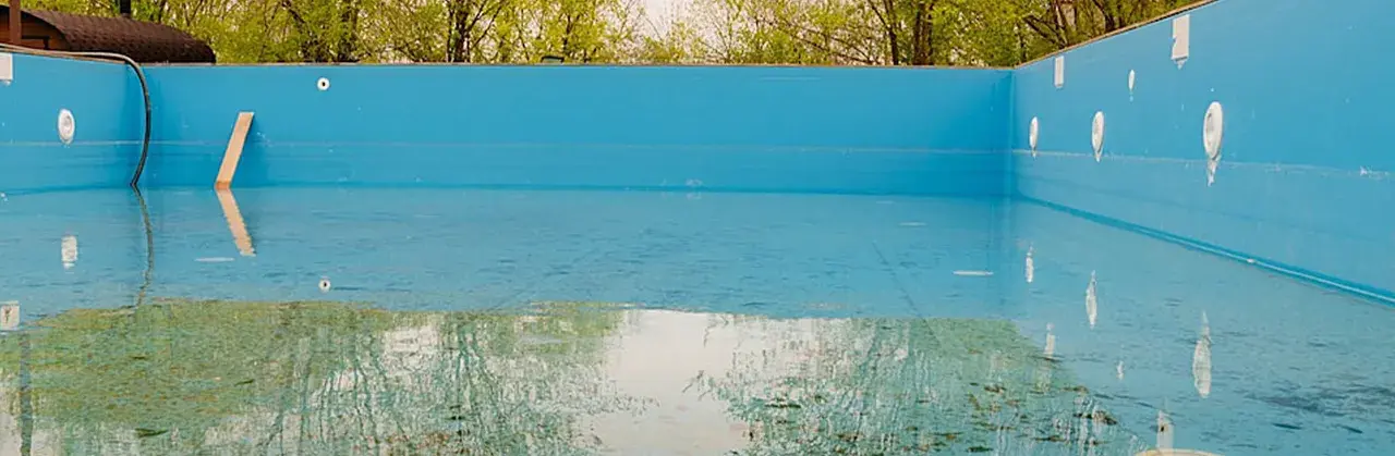 La piscina in inverno va svuotata. L'acqua residua riflette il verde degli alberi e il cielo, mentre il fondo mostra i segni del tempo.