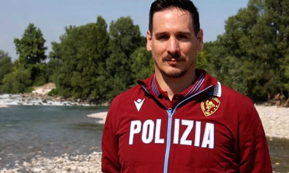 Federico Vanelli, con indosso una giacca della Polizia, posa vicino a un fiume con alberi sullo sfondo.