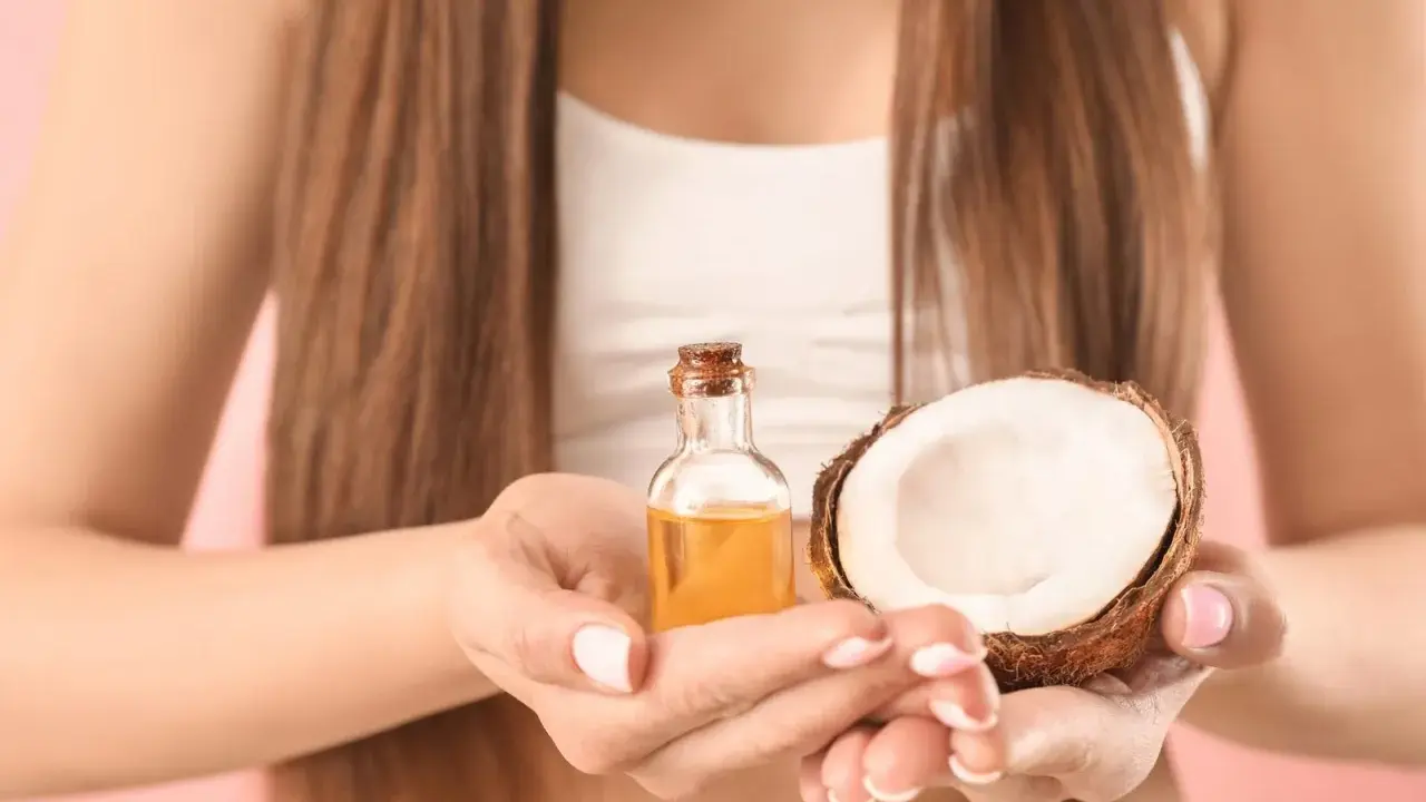 Mani che tengono una boccetta di olio di cocco e mezzo cocco. Ideale per proteggere i capelli dopo la piscina.