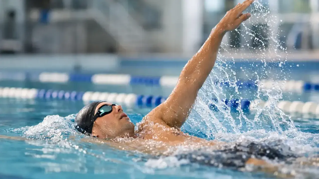 Atleta in piscina esegue la nuotata a **scheda dorso**, con il braccio destro sollevato e l'acqua che schizza.
