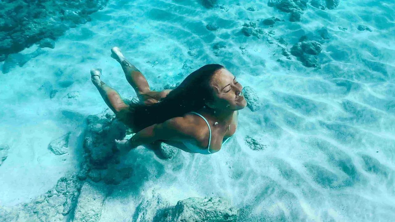 Donna nuota sott'acqua, superando la paura delle acque profonde. La luce filtra attraverso l'acqua cristallina, illuminando la sabbia e le rocce sul fondale.