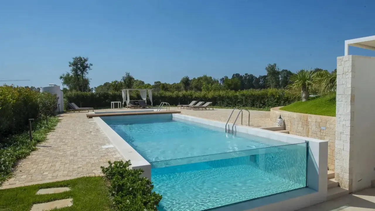 Piscina fuori terra su terreno in pendenza con pareti in vetro, circondata da un giardino curato e un'area relax con lettini e gazebo.
