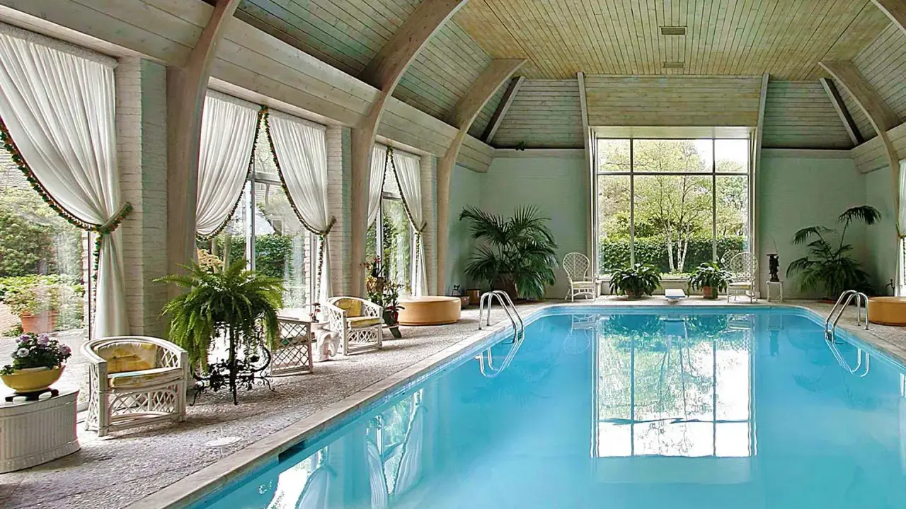 Piscina interna con acqua azzurra, sedie in vimini, piante e grandi finestre. Uno skimmer piscina cos'è? Serve a raccogliere detriti galleggianti.