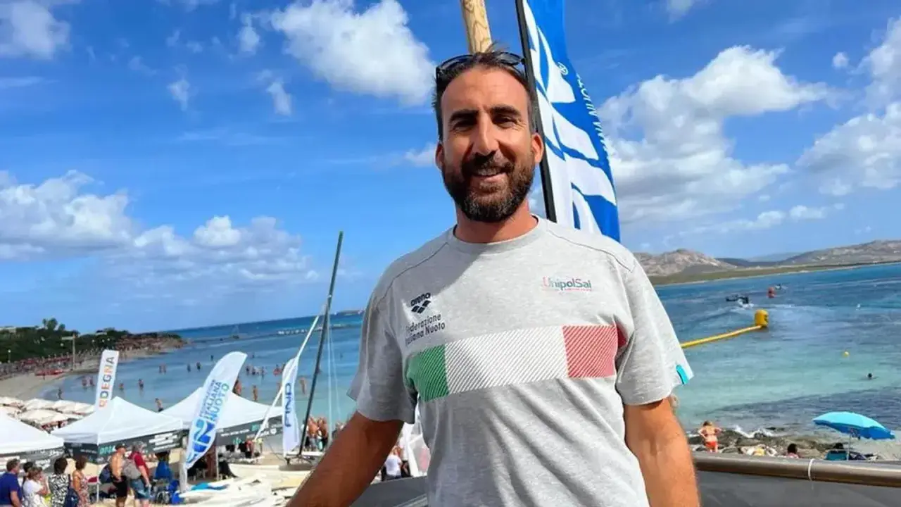 Fabrizio Antonelli sorride in spiaggia, con la bandiera italiana sul petto, pronto per un evento di nuoto.