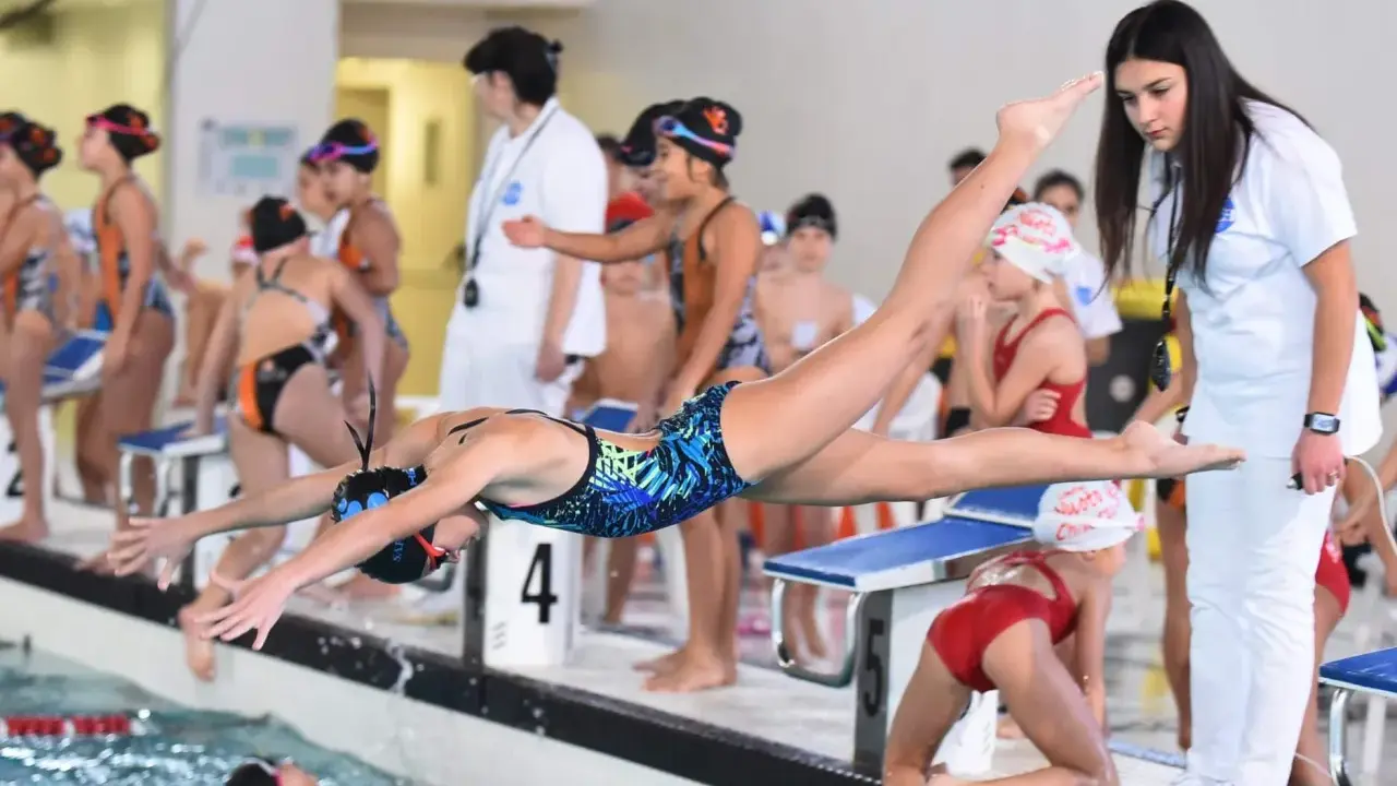 Axana Merckx, giovane nuotatrice, si tuffa in piscina con agilità. Allenatrice osserva la gara.
