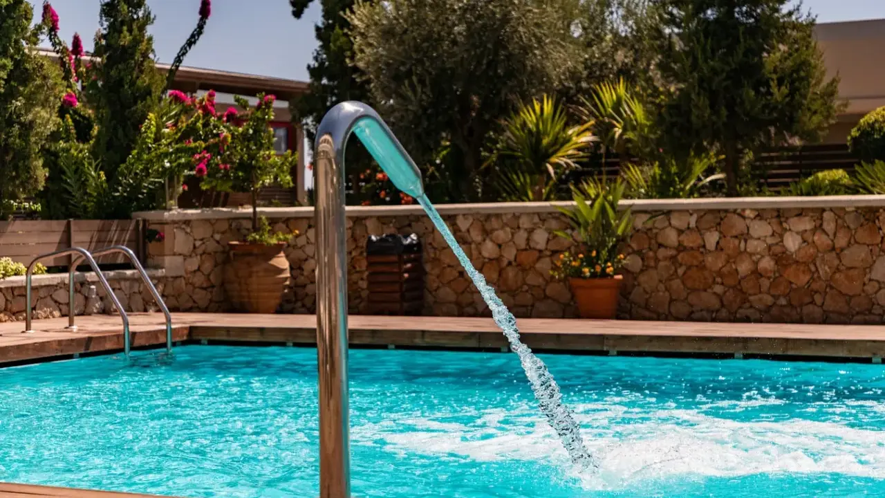 Un getto d'acqua da una fontana metallica riempie la piscina azzurra, circondata da legno e vegetazione lussureggiante.
