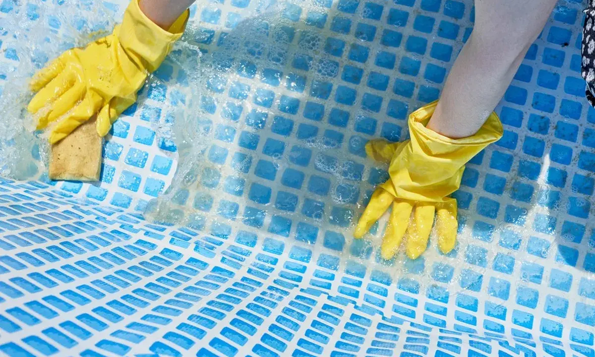Mani con guanti gialli puliscono il fondo di una piscina con una spugna, mostrando come togliere calcare dalla piscina.