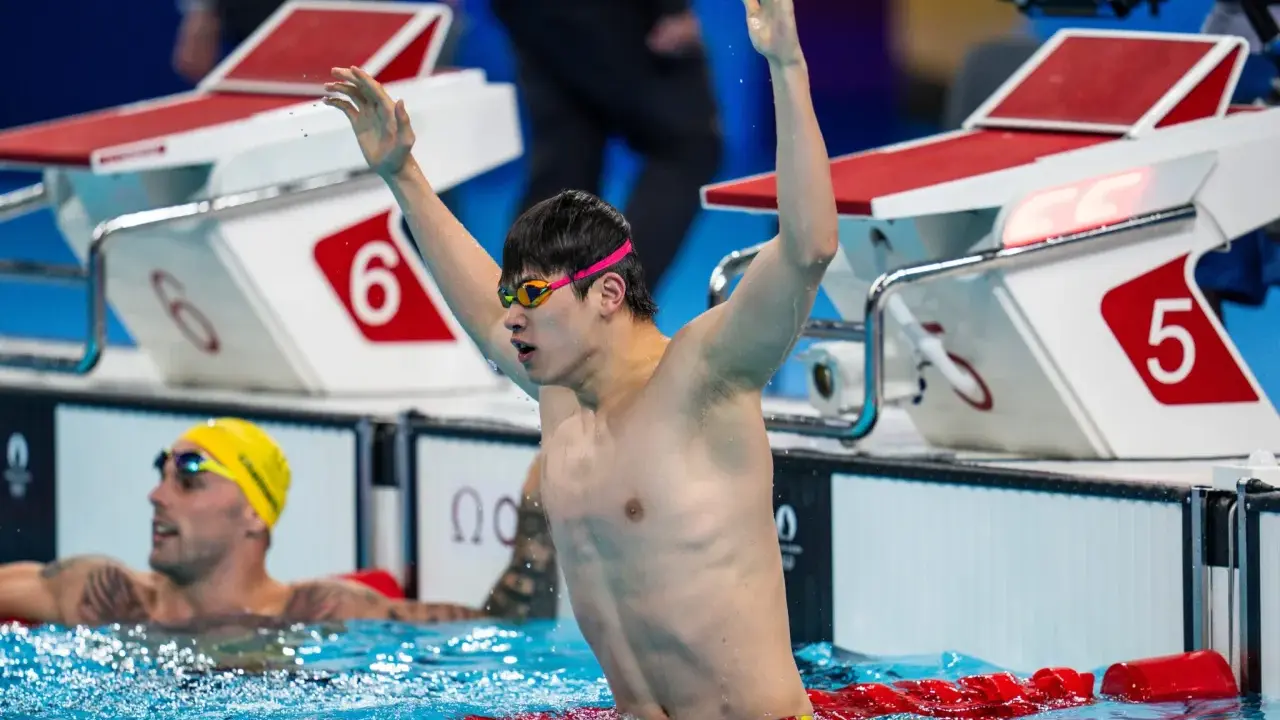 Nuotatore con braccia alzate, festeggia la vittoria. Pan Zhanle trionfa in piscina, circondato da avversari e blocchi di partenza.
