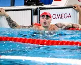 Adam Peaty, con cuffia rossa e occhialini, emerge dall'acqua della piscina, aggrappato al bordo.