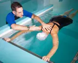 Fisioterapista assiste donna in piscina per ginnastica in acqua per mal di schiena. Lei galleggia con un supporto, lui la guida.
