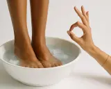 Piedi immersi in una ciotola con acqua fredda e cubetti di ghiaccio, un gesto con la mano indica che il **shock termico acqua fredda** è perfetto per un sollievo immediato.