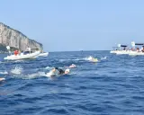 Nuotatori in gara tra Capri e Napoli, con barche di supporto e la costa rocciosa sullo sfondo.