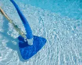 Pulizia di una piscina sporca con aspiratore blu e manico in legno. L'acqua limpida riflette la luce creando un effetto scintillante.