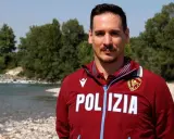 Federico Vanelli, con indosso una giacca della Polizia, posa vicino a un fiume con alberi sullo sfondo.