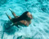 Donna nuota sott'acqua, superando la paura delle acque profonde. La luce filtra attraverso l'acqua cristallina, illuminando la sabbia e le rocce sul fondale.