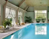 Piscina interna con acqua azzurra, sedie in vimini, piante e grandi finestre. Uno skimmer piscina cos'è? Serve a raccogliere detriti galleggianti.