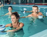 Persone sorridenti fanno sport in piscina con attrezzi galleggianti per un allenamento acquatico tonificante.