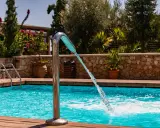 Un getto d'acqua da una fontana metallica riempie la piscina azzurra, circondata da legno e vegetazione lussureggiante.