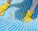 Mani con guanti gialli puliscono il fondo di una piscina con una spugna, mostrando come togliere calcare dalla piscina.