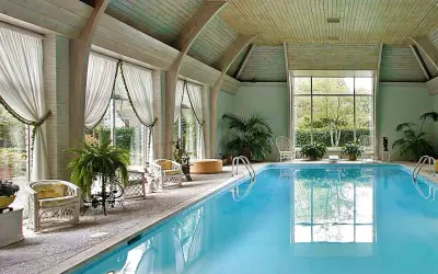 Piscina interna con acqua azzurra, sedie in vimini, piante e grandi finestre. Uno skimmer piscina cos'è? Serve a raccogliere detriti galleggianti.