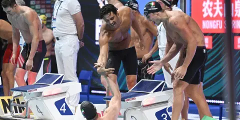 Atleti italiani festeggiano dopo la staffetta 4x100. Un nuotatore porge la mano a un compagno, gioia e sudore bagnano la corsia.