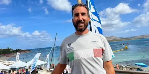 Fabrizio Antonelli sorride in spiaggia, con la bandiera italiana sul petto, pronto per un evento di nuoto.