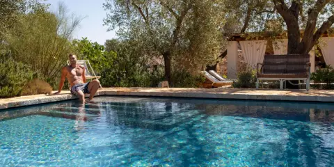 Uomo seduto sul bordo di una piscina con acqua salata, circondata da ulivi e vegetazione mediterranea.
