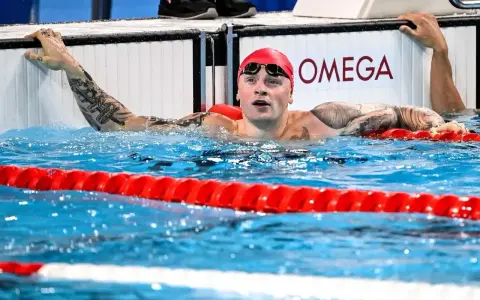 Adam Peaty, con cuffia rossa e occhialini, emerge dall'acqua della piscina, aggrappato al bordo.