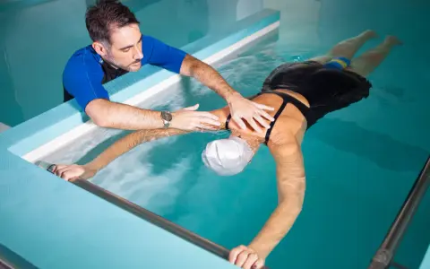 Fisioterapista assiste donna in piscina per ginnastica in acqua per mal di schiena. Lei galleggia con un supporto, lui la guida.