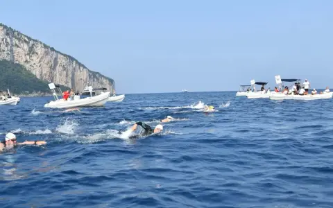 Nuotatori in gara tra Capri e Napoli, con barche di supporto e la costa rocciosa sullo sfondo.