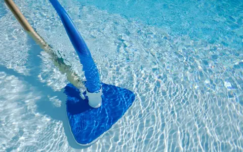 Pulizia di una piscina sporca con aspiratore blu e manico in legno. L'acqua limpida riflette la luce creando un effetto scintillante.