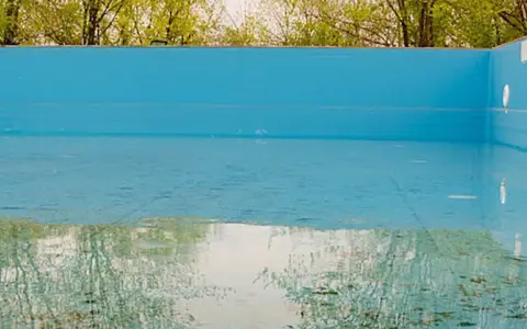 La piscina in inverno va svuotata. L'acqua residua riflette il verde degli alberi e il cielo, mentre il fondo mostra i segni del tempo.
