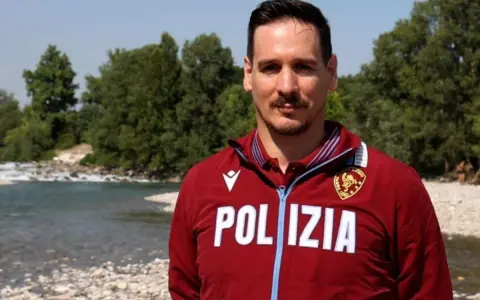 Federico Vanelli, con indosso una giacca della Polizia, posa vicino a un fiume con alberi sullo sfondo.
