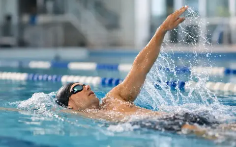 Atleta in piscina esegue la nuotata a **scheda dorso**, con il braccio destro sollevato e l'acqua che schizza.