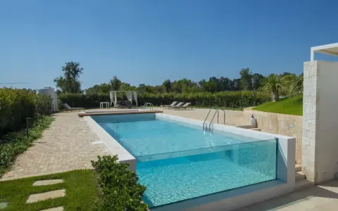 Piscina fuori terra su terreno in pendenza con pareti in vetro, circondata da un giardino curato e un'area relax con lettini e gazebo.