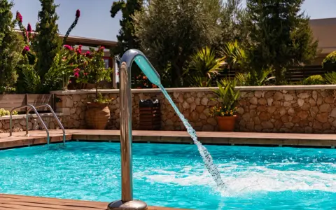 Un getto d'acqua da una fontana metallica riempie la piscina azzurra, circondata da legno e vegetazione lussureggiante.