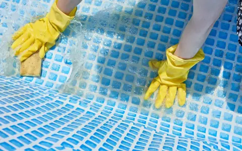 Mani con guanti gialli puliscono il fondo di una piscina con una spugna, mostrando come togliere calcare dalla piscina.