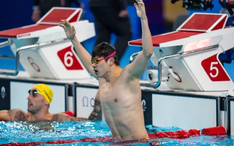 Nuotatore con braccia alzate, festeggia la vittoria. Pan Zhanle trionfa in piscina, circondato da avversari e blocchi di partenza.