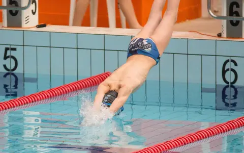 Un atleta si tuffa in piscina, mostrando la sua massa muscolare sviluppata grazie al nuoto. L'acqua schizza mentre entra in azione.
