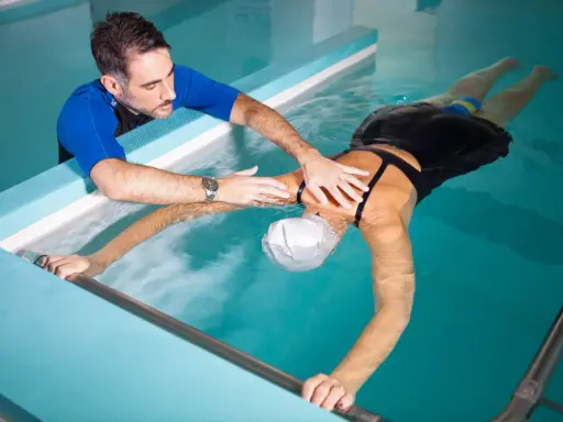 Fisioterapista assiste donna in piscina per ginnastica in acqua per mal di schiena. Lei galleggia con un supporto, lui la guida.