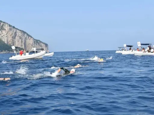 Nuotatori in gara tra Capri e Napoli, con barche di supporto e la costa rocciosa sullo sfondo.