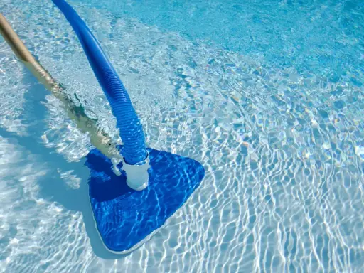 Pulizia di una piscina sporca con aspiratore blu e manico in legno. L'acqua limpida riflette la luce creando un effetto scintillante.