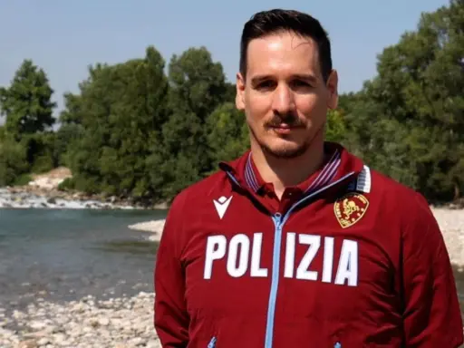 Federico Vanelli, con indosso una giacca della Polizia, posa vicino a un fiume con alberi sullo sfondo.