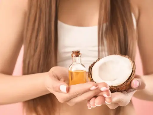 Mani che tengono una boccetta di olio di cocco e mezzo cocco. Ideale per proteggere i capelli dopo la piscina.
